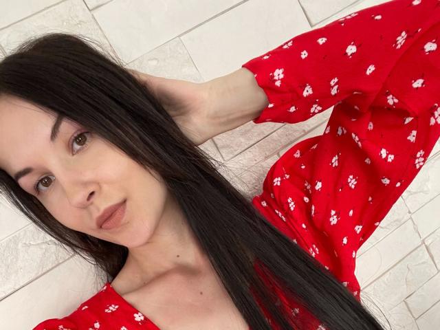 JuliaSexxx-hot has long black hair, light skin, and brown eyes. She's wearing a red dress with white floral patterns, lying against a white tiled wall. Her right hand is in her hair, and she's looking slightly to the right with a neutral expression. The dress's fabric looks soft and slightly textured. The tiles have a subtle, patterned design. The image is a close-up, focusing on her face and upper body. The lighting is even, highlighting her facial features and dress.