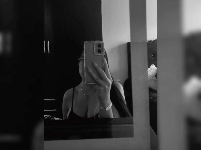 Black-and-white photo of MiaKhalipso taking a mirror selfie. She holds a phone with a studded case, partially covering her face. Her right hand is visible, wearing a wristwatch. She's wearing a strapless top, showing cleavage. The mirror has a dark frame, and a blurred object is on the right. The phone's camera is centered, and her face is obscured. The image has a high-contrast, gritty texture.