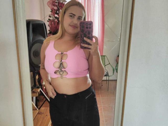 LadyMiss with blonde hair, pink crop top with cut-out circles, showing her big tits, black skirt, holding phone, mirror selfie, smiling, pink curtains, wooden floor, speaker behind.