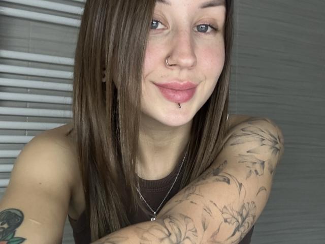 VikiKissX with brown hair, nose ring, lip piercing, and tattooed arm, wearing a brown tank top, smiling softly. Her tits are small, and she's cute.