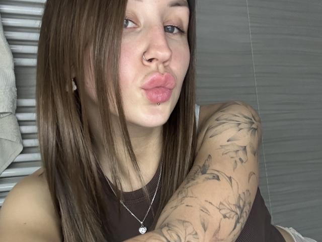 VikiKissX with brown hair, nose piercing, and lip piercing, pouting with pink lips, tattooed arm, brown tank top, and necklace, in a bathroom.