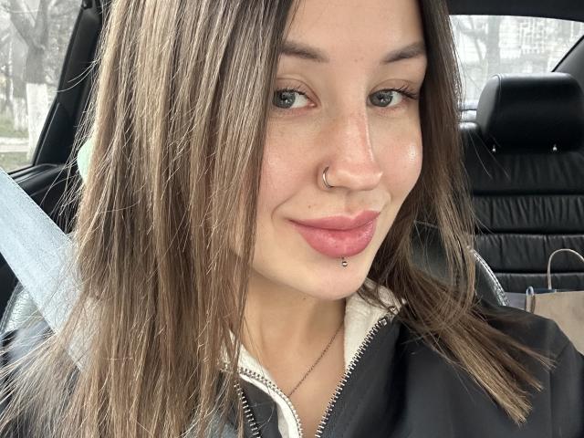 VikiKissX has brown hair, a nose ring, and a lip piercing with a small ball. She's wearing a black jacket and a white zip-up shirt, smiling at the camera.