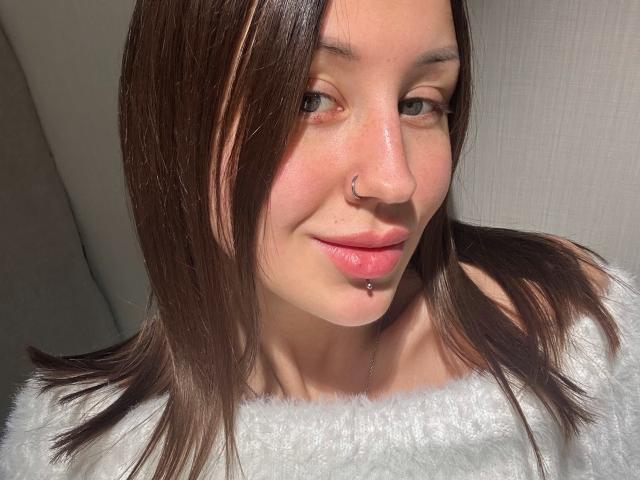 VikiKissX has brown hair, a nose ring, and a lip piercing, wearing a white fuzzy top. She's smiling with sunlit skin and green eyes.