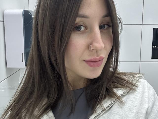 VikiKissX has brown hair, a nose ring, and pink lips. She's wearing a white zip-up and a gray shirt. Her eyes are blue-green. She's in a bathroom.