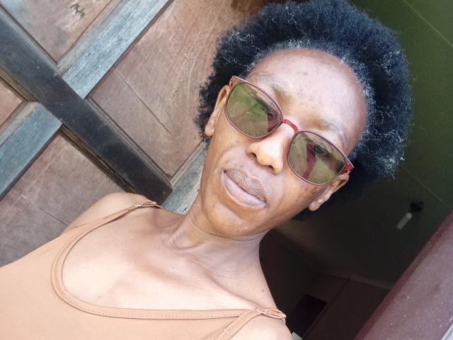 LindaTave has dark brown skin, short curly black hair, red-framed sunglasses, and a tan tank top, showing a slightly puffy lower lip. Her face is slightly wrinkled.