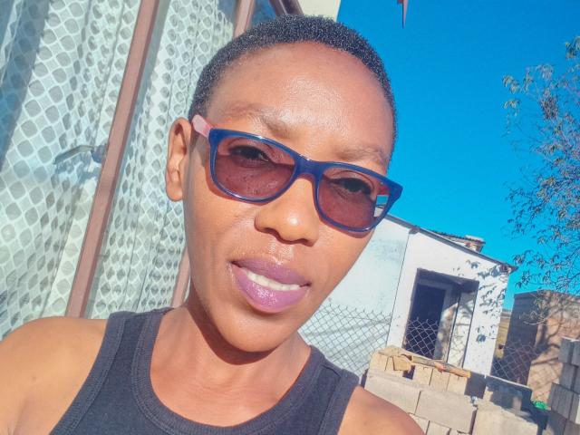 LindaTave with short black hair, blue sunglasses, and purple lipstick, wearing a black tank top, smiling outside under a bright blue sky.