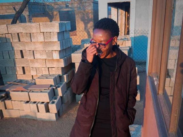 LindaTave with dark skin, short curly hair, wearing red glasses, black shirt, and brown jacket, standing by stacked concrete bricks, covering her mouth with one hand.