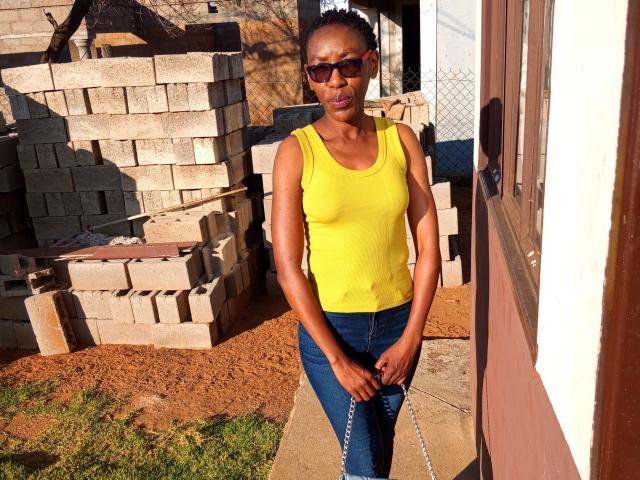 LindaTave with dark skin, wearing a yellow tank top, blue jeans, and red sunglasses, standing in a sunlit yard with a brick wall and a house on the right.