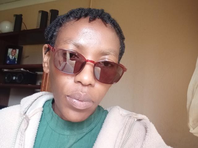 LindaTave with short black curly hair, wearing red sunglasses, green turtleneck, and beige zip-up jacket, has a slightly shiny forehead and a neutral expression.