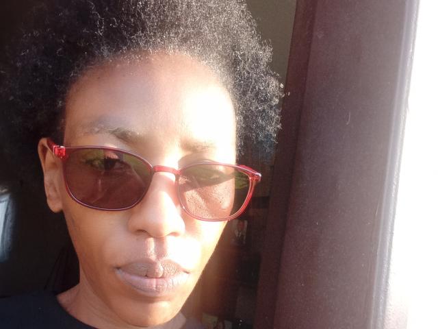 LindaTave with dark curly hair, brown skin, red sunglasses, and a serious expression, standing in sunlight next to a brown curtain. Her ass is not visible.