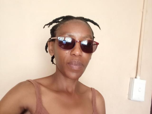 LindaTave with dark brown skin, small boobs, and braids, wearing red sunglasses, and a brown tank top, looking serious, against a beige wall.
