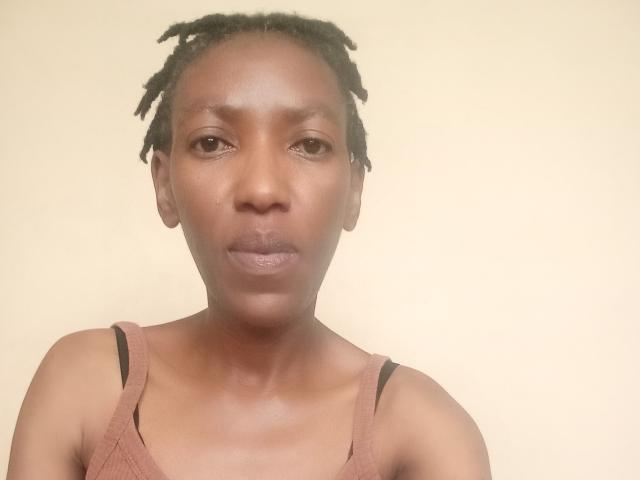 LindaTave with dark brown skin, small black bra straps, and short black dreadlocks, looking straight at the camera with a neutral expression.