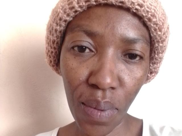LindaTave with a brown crochet hat, dark brown skin, small nose, slightly parted lips, and subtle freckles on her face, wearing a white shirt.