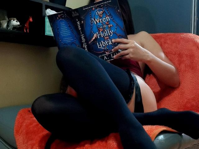 SugarSpiceXo is reading a book, wearing black thigh-highs and black panties, with her legs crossed on a red chair, exposing her bare ass. Her face is hidden by the book.
