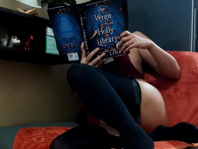 SugarSpiceXo with books covering tits, wearing black thigh-highs and red lingerie, lying on a red towel, legs up, reading "The Winter of Holy Library" in dim lighting.