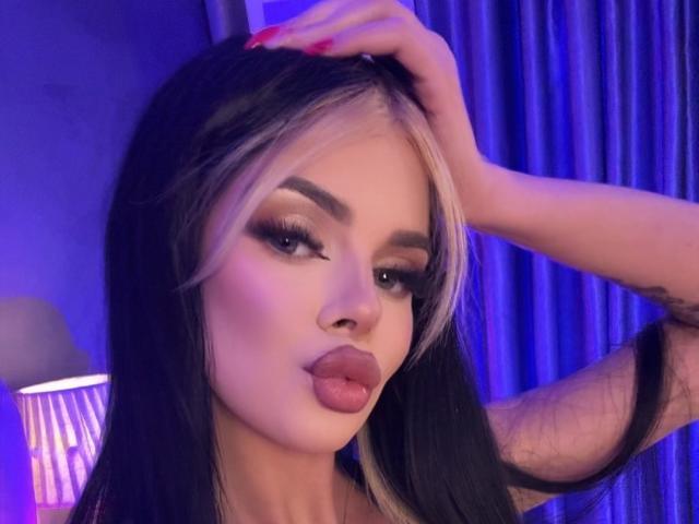 IvyRaven with black and blonde hair, pouty lips, and red nails, hand in hair, blue and purple lighting, eyeliner, and full makeup, looking seductive.