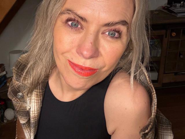 MiaHase-hot has gray eyes, red lipstick, and a slight smile. She's wearing a plaid shirt over a black top, with her right shoulder exposed. She's a middle-aged woman.