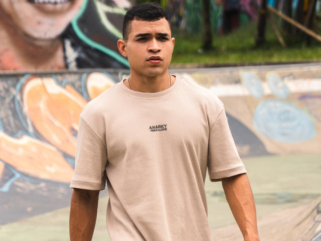 GersonZT has a medium build, short dark hair, and is wearing a beige t-shirt. His face is serious, with slightly parted lips, in an outdoor urban setting.