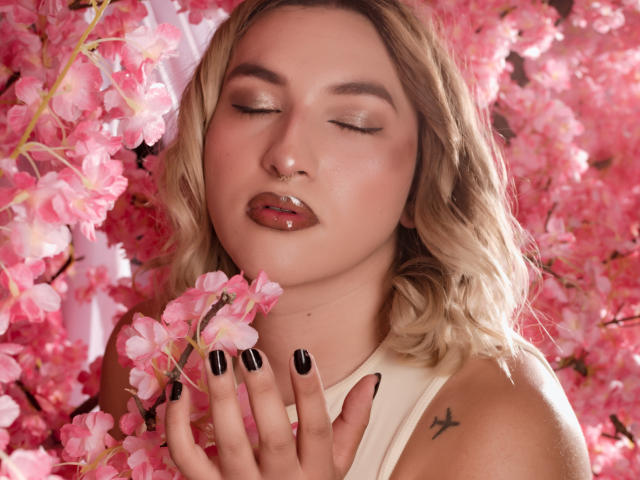 TommyTS met roze kersenbloesems, ogen gesloten, glanzende roze lippenstift, zwarte nagellak, blond golvend haar, kleine tatoeage op de rechter schouder, wit mouwloos topje, zachte roze en roze achtergrond..