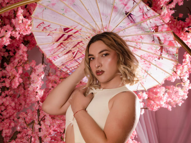 TommyTS with blonde hair, wearing a white sleeveless top, holding a pink cherry blossom umbrella, pink cherry blossoms in the background, looking at the camera with red lipstick.