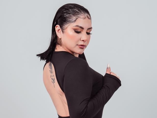 AntonellaFerreer has wet black hair, wearing a tight black long-sleeve top, showing her tattooed arm, and touching her small tits with a slight smile, against a plain background.
