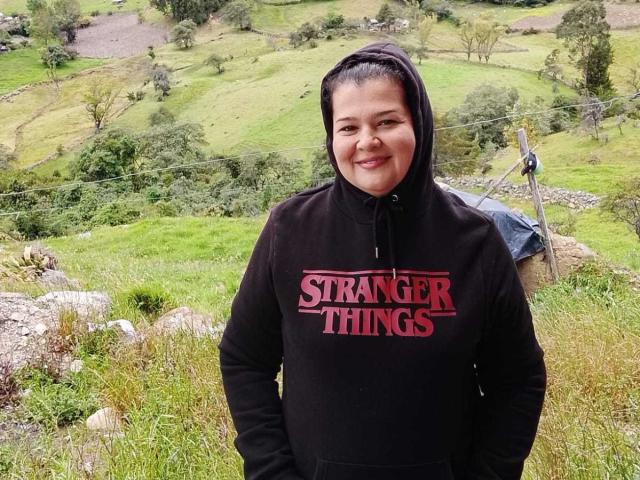 AdrianaPrettie, chubby, smiling, wearing a black hoodie with "STRANGER THINGS" in red, standing in a grassy, rocky area, hands in pockets, dark hair in a ponytail.