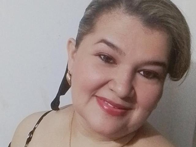 AdrianaPrettie with short brown hair, smiling, glossy pink lips, small nose, black strap top, gold earrings, and a gold necklace. Her tits are small, she's cute.