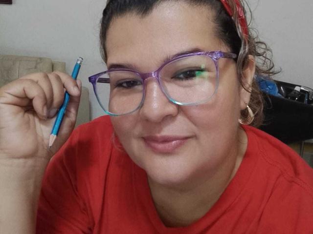 AdrianaPrettie with purple and green gradient glasses, holding a blue pencil, wearing a red shirt, and a red headband, smiling slightly, with hoop earrings.