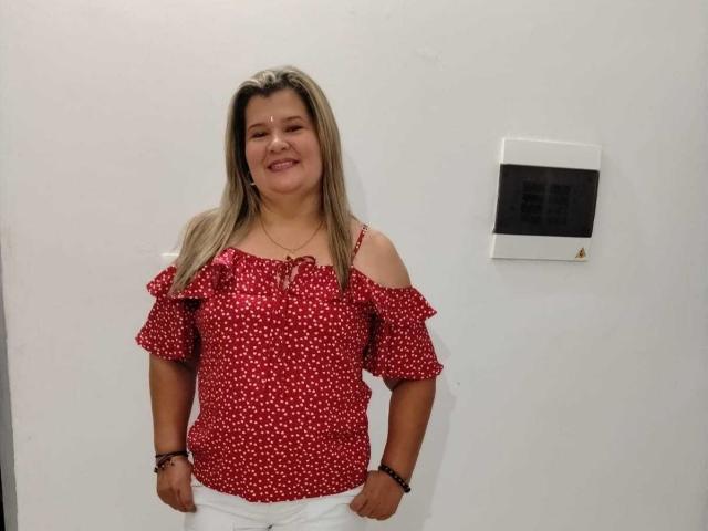 AdrianaPrettie has blonde hair, red off-shoulder polka dot top, white pants, smiling, standing, wall thermostat on right, wearing bracelet, looking relaxed.