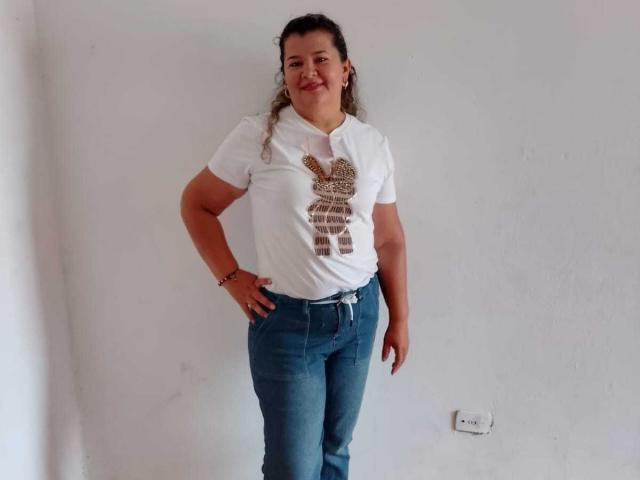 AdrianaPrettie stands with hands on hips, wearing a white tee with a leopard print design, blue jeans, and a smile. She's got curly brown hair and a tan skin tone.