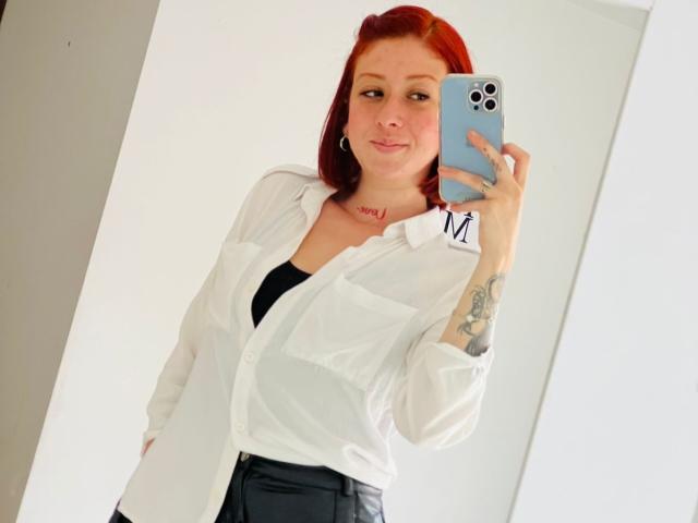 Valeriaaf with red hair, white button-up shirt, black top, black pants, holding blue phone, tattoo on right arm, smiling, standing in front of mirror.
