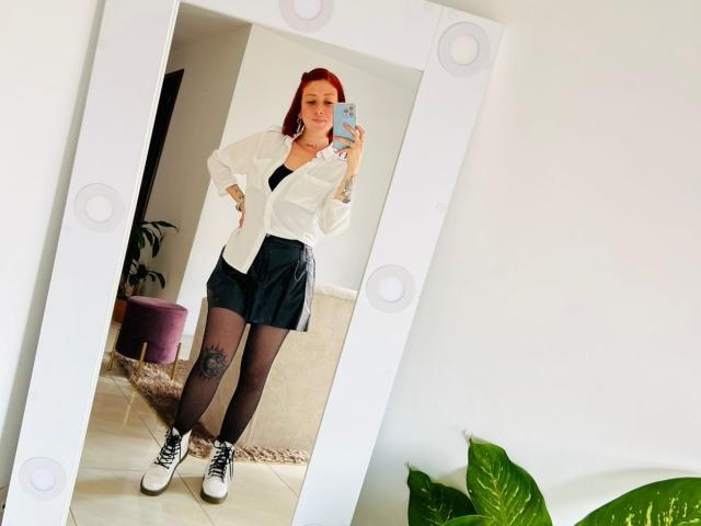 Valeriaaf with red hair, white shirt, black skirt, black tights, white sneakers, standing in front of mirror, holding phone, tattoo on left thigh, green plant in bottom right.