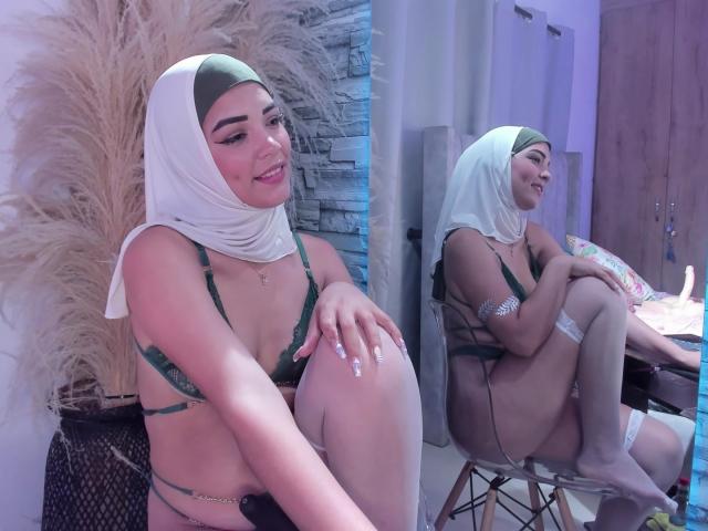 AliceKayleigh, wearing a green bra and white hijab, sits with her knees up, showing her small tits and cute smile. Reflection in mirror shows her smiling too.