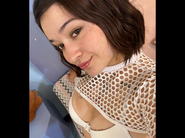 AnikaFleur, con cabello castaño corto, vistiendo una blusa blanca de malla que revela sus pechos y un sostén blanco, sonriendo ligeramente, con el nose piercing, en un selfie..