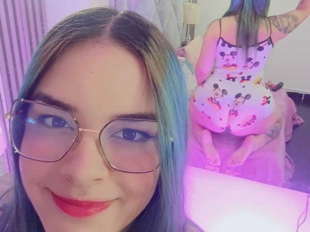 AuroraSantorini with blue-green hair, glasses, and red lipstick, wearing a Mickey Mouse print onesie, showing her curvy ass and tattoos, kneeling on a bed, smiling at the camera.