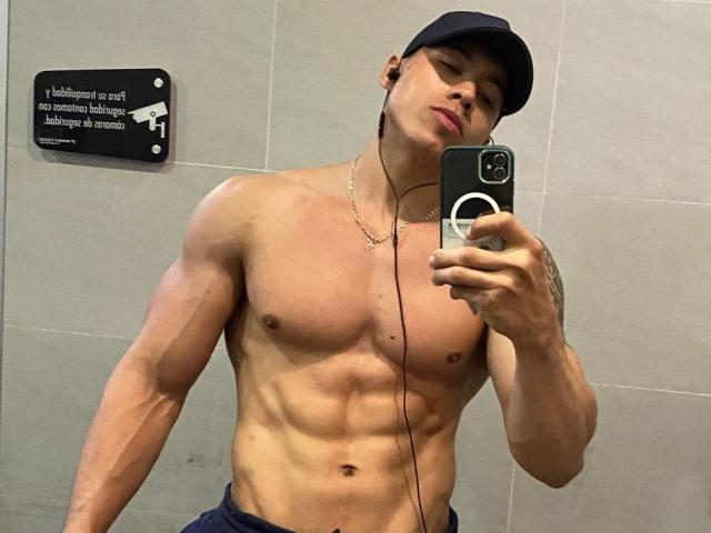AlecAndAndrus, shirtless with a muscular, defined, tan upper body, wearing a black cap, holding a phone, earbuds in, mirror selfie, necklace, and blue jeans.