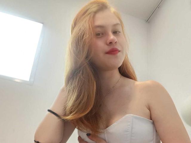 TaylorCarpert with long red hair, wearing a white strapless top, has small tits, light skin, red lipstick, black bracelet, and a slight smile, looking at the camera.