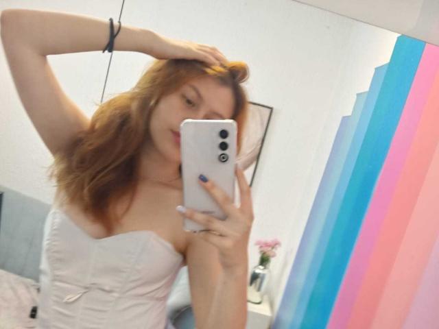 TaylorCarpert with red hair, wearing a white strapless dress, taking a mirror selfie with her left hand in her hair, showing her tits and holding a white phone.