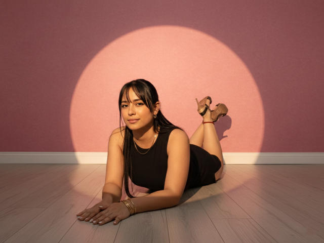 GahiaBelucci is lying on a wooden floor, wearing a black dress, with her ass up and heels on, a golden bracelet, and a pink sunshadow on a pink wall.