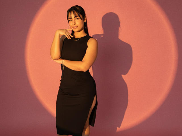 GahiaBelucci in a tight black dress with side slits, standing with one hand near her chin, shadow on pink wall, sunlight highlighting her curves and medium tits.