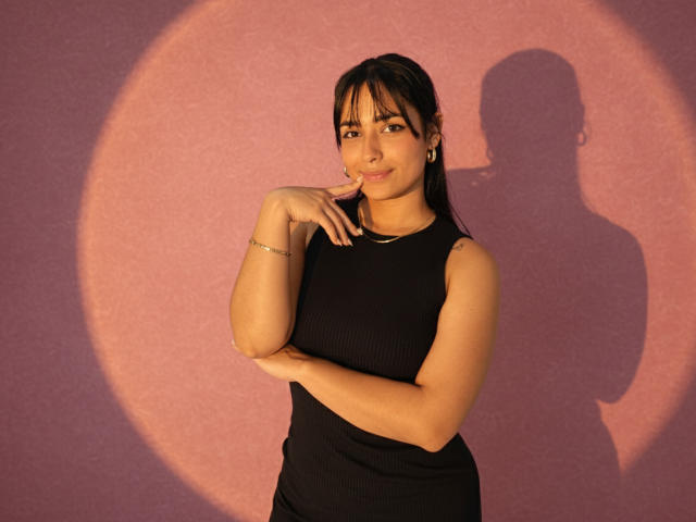 GahiaBelucci with dark hair, wearing a black ribbed sleeveless top, has a bracelet on her right wrist, and is touching her chin with a slight smile. Her shadow is on the wall.