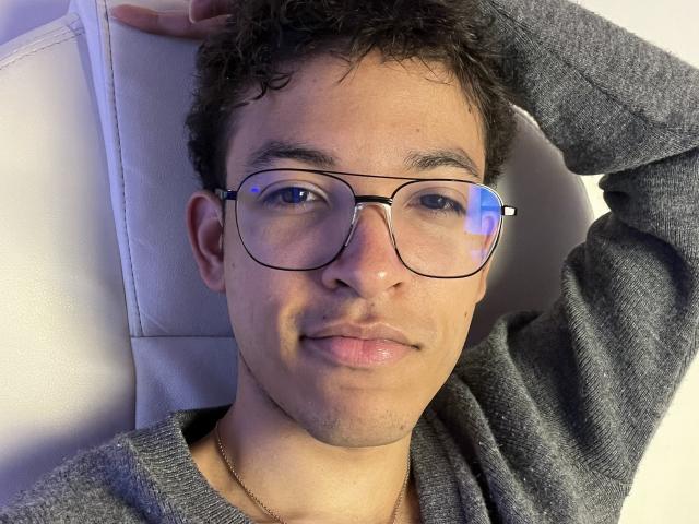 MiloJhones with curly dark hair, wearing glasses and a gray sweater, has a slight smile and a gold necklace, lying back with one hand on his head.