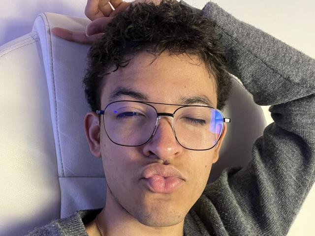MiloJhones with curly dark hair, wearing glasses and a grey sweater, pouting his lips, has a gold necklace, and is resting his head on a white chair.