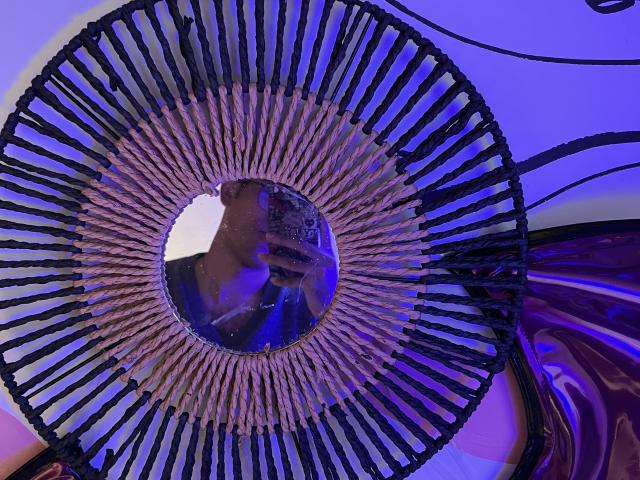 MiloJhones' face in a mirror, surrounded by a circular black and white striped pattern, with blue and purple lighting. Reflection shows MiloJhones' upper body and phone.