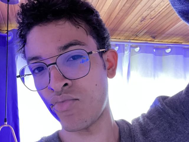 MiloJhones with curly black hair, glasses, and a small pipe in his mouth, wearing a grey sweater, in a selfie with a wooden ceiling. His face is slightly pouty.