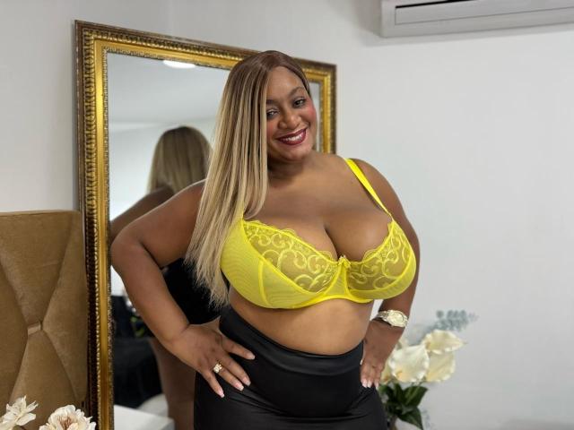 NahomiAnders with large yellow lace bra showing her big tits, black leather skirt, and smiling. Blonde hair, tan skin, white wristwatch, gold mirror, white wall, and air conditioner.