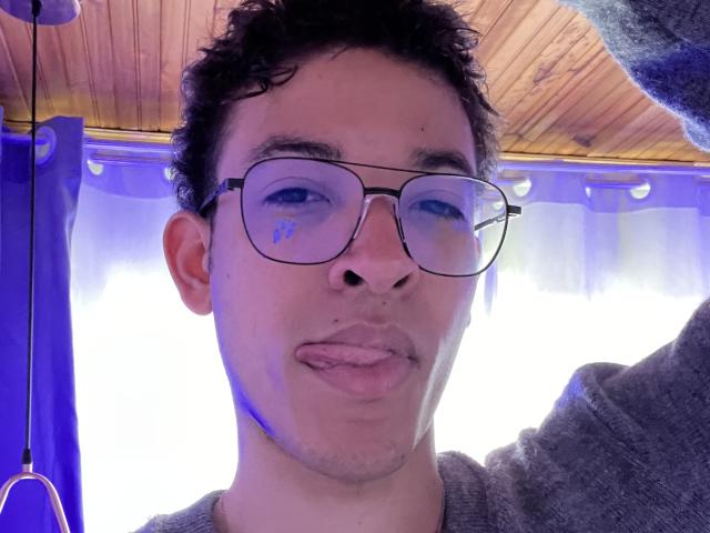 MiloJhones with glasses, sticking out tongue, short curly dark hair, light skin, grey sweater, wood ceiling, blue curtain, selfie, slight blue reflection in glasses.