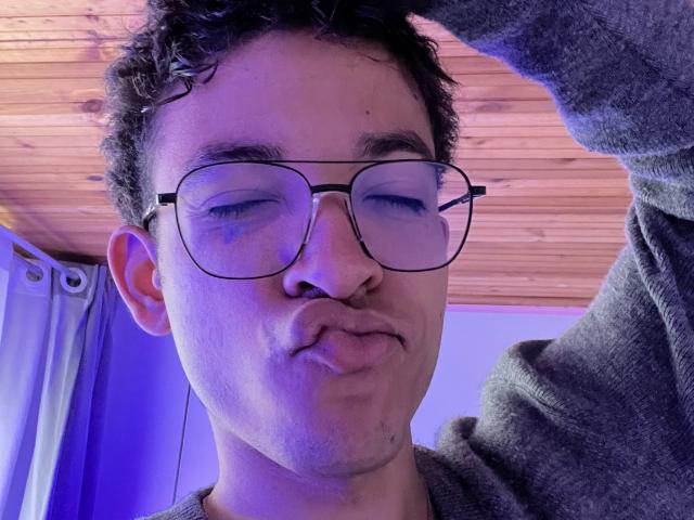 MiloJhones, with curly dark hair, wearing large glasses and a grey hoodie, puffs out his cheeks in a selfie, wood ceiling and blue curtain in the background.