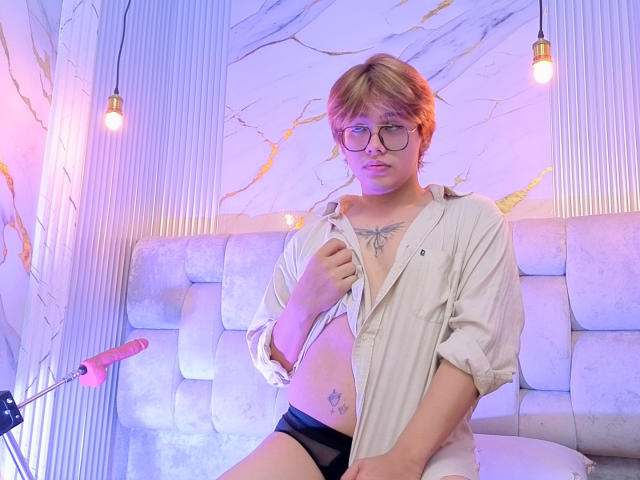 BenjiVampire with glasses, blonde hair, open shirt, black underwear, chest tattoo, and thigh tattoo, sitting on white cushioned bench, pink dildo on stand, purple and blue lights.