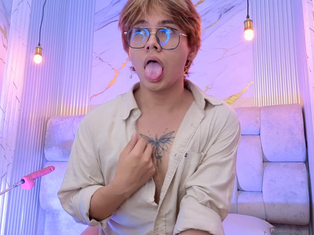 BenjiVampire with short blonde hair, glasses, and tongue out, wearing a beige shirt, pointing at a tattoo on their chest, with a pink dildo on the left.